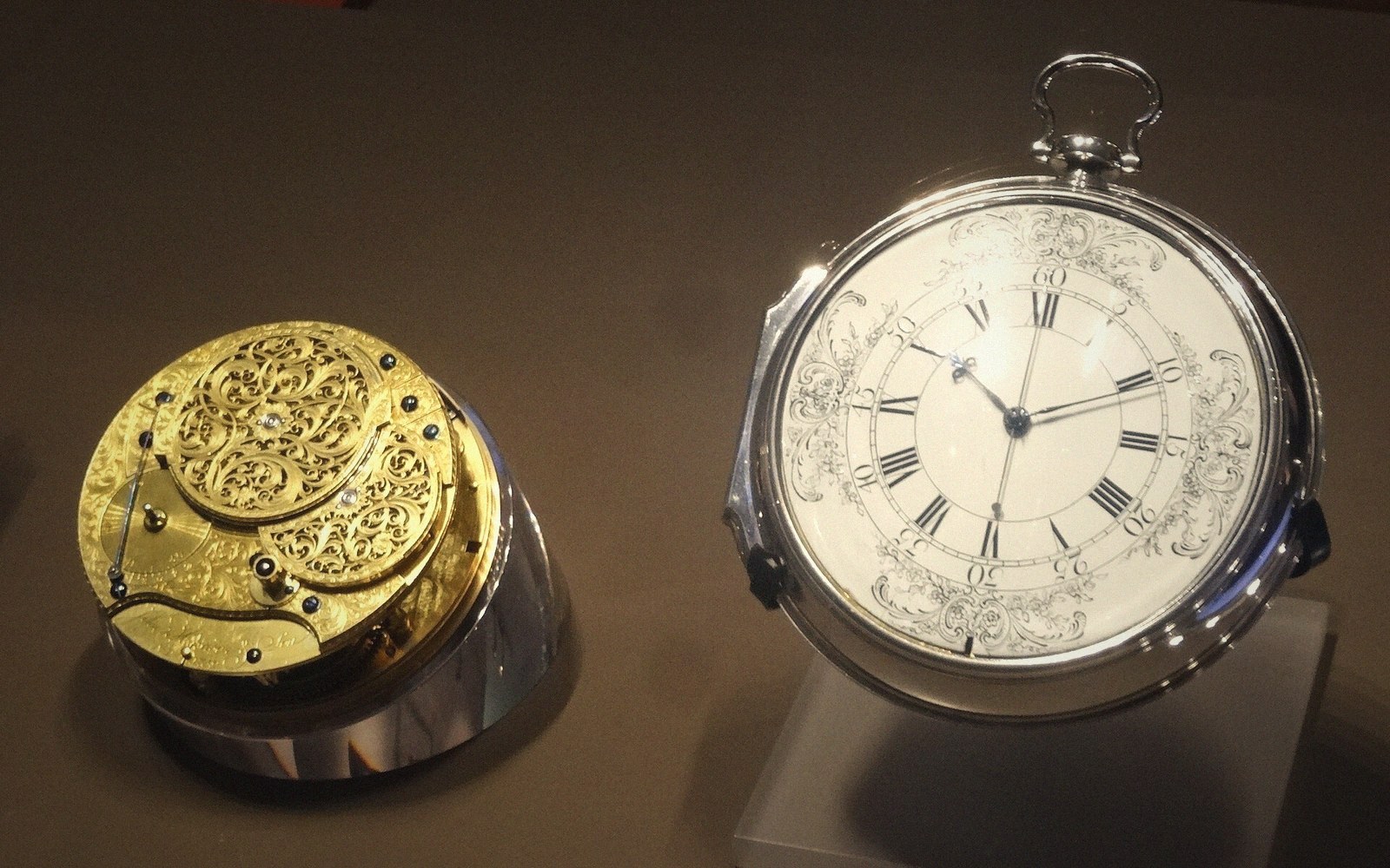 A large ornate pocket watch movement displayed under glass in a museum case, dial removed to show the mechanism, controlled gallery lighting